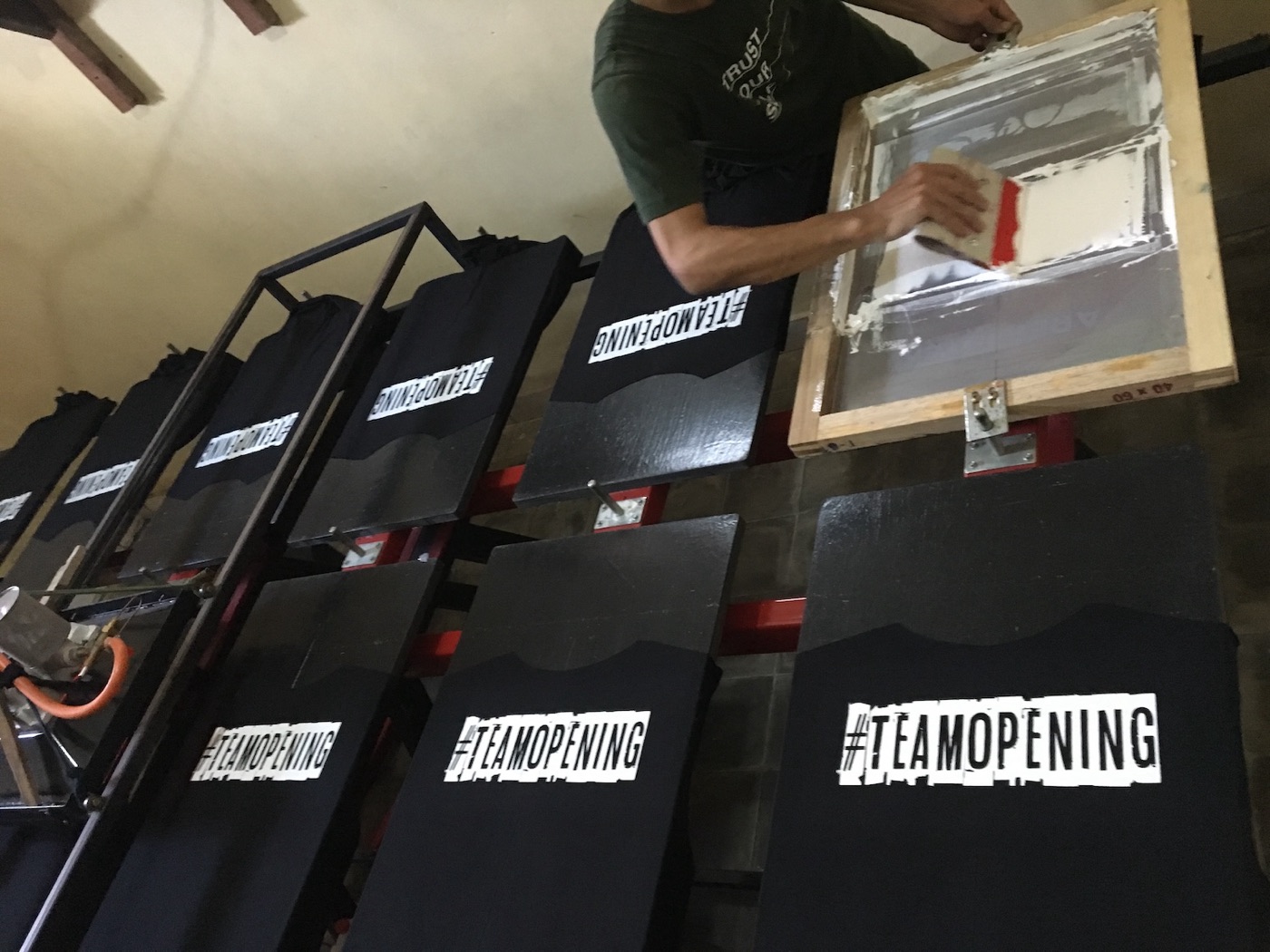 A person pulling a screen-print squeegee over black t-shirts racked in rows.